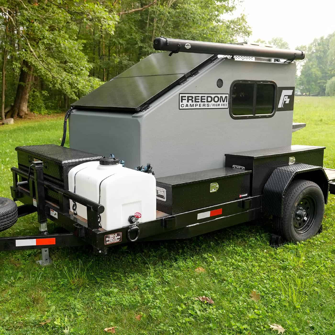 Freedom Camper on a black utility trailer with a front water tank, side toolbox, and roof-mounted awning