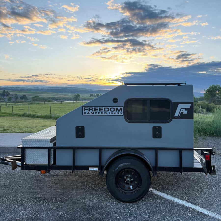 Gray Freedom Camper bolted to a black utility trailer at sunset