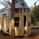 Small wooden tiny house with large windows and metal roof in a wooded area.