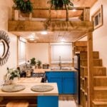 Interior of the Goa tiny house with bright blue cabinets, a full-size range, a butcher-block peninsula, a sleeping loft above, and an open storage staircase