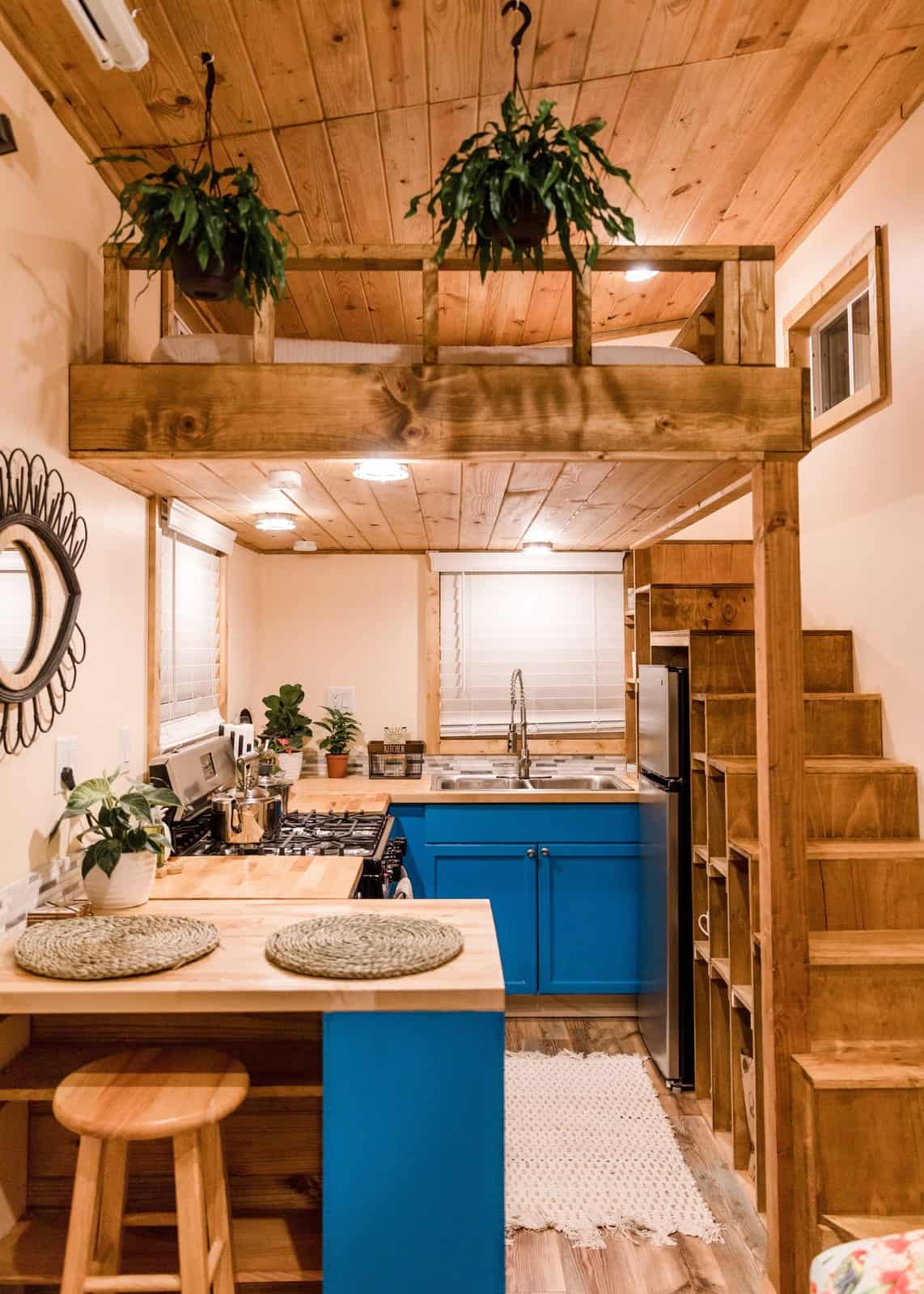 Interior of the Goa tiny house with bright blue cabinets, a full-size range, a butcher-block peninsula, a sleeping loft above, and an open storage staircase