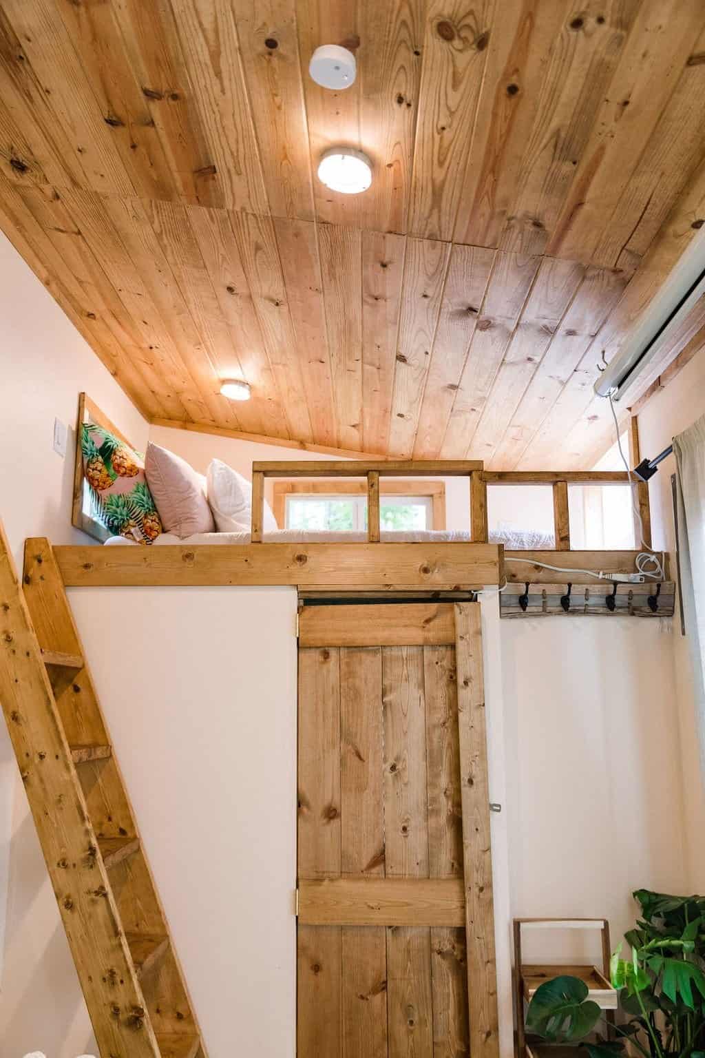 Queen sleeping loft in the Goa tiny house reached by ladder, with a pine ceiling and a barn door below