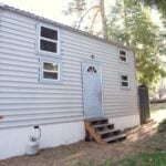 White-sided tiny house with a pale-blue front door and arched fan window, blue-trimmed windows, wooden entry steps and a propane tank, set among pines at Tiny House Block in Mt. Laguna, California