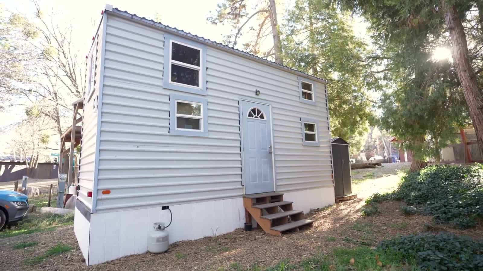 White-sided tiny house with a pale-blue front door and arched fan window, blue-trimmed windows, wooden entry steps and a propane tank, set among pines at Tiny House Block in Mt. Laguna, California