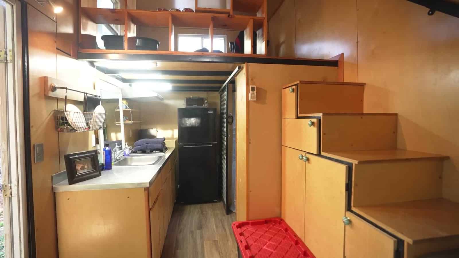 Galley kitchen in a tiny house with a stainless sink, black refrigerator, open loft shelving above, and plywood storage stairs with glass knobs leading to the second loft