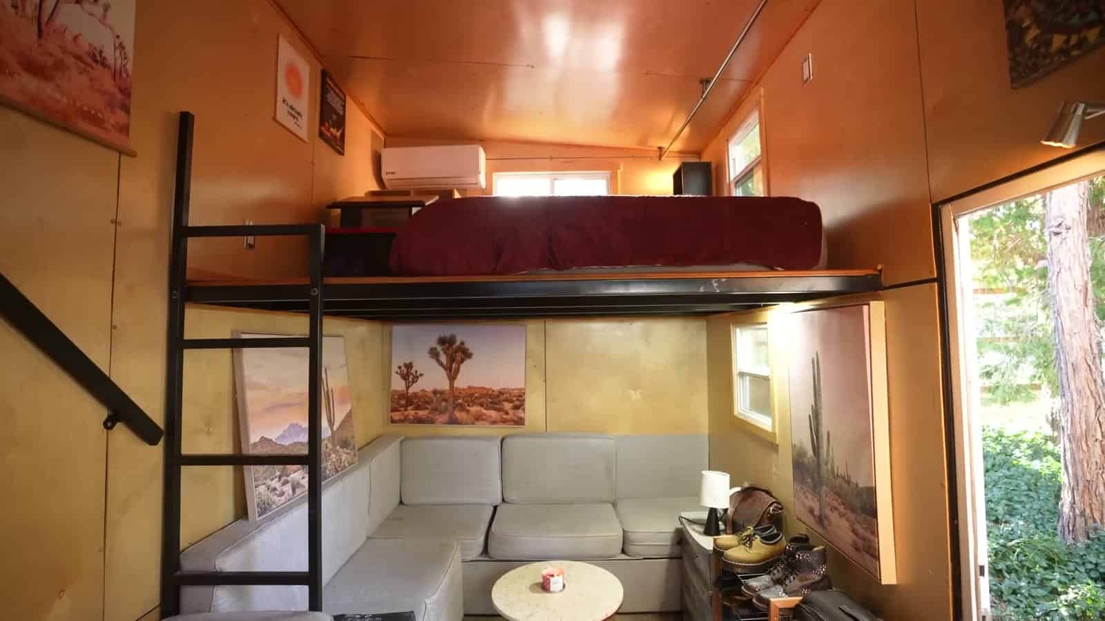 Tiny house living room with a pale sectional sofa, round table, desert prints, and a red-topped loft bed overhead reached by a black metal ladder