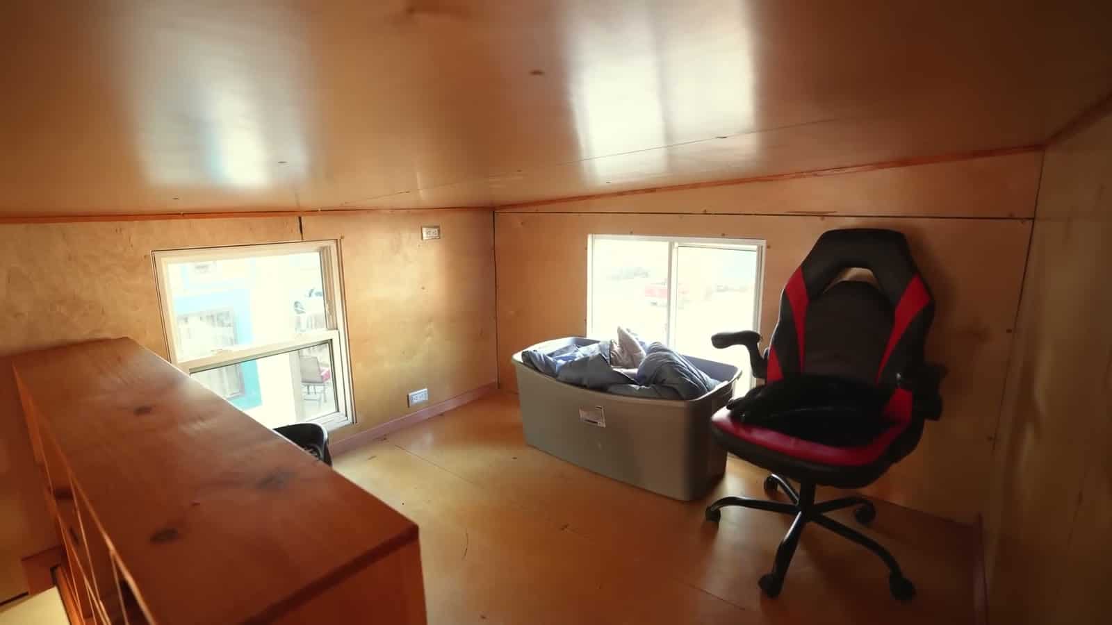 Second loft in a tiny house used as an office, with a long wood desk ledge, gaming chair, storage bin and two windows looking into the trees