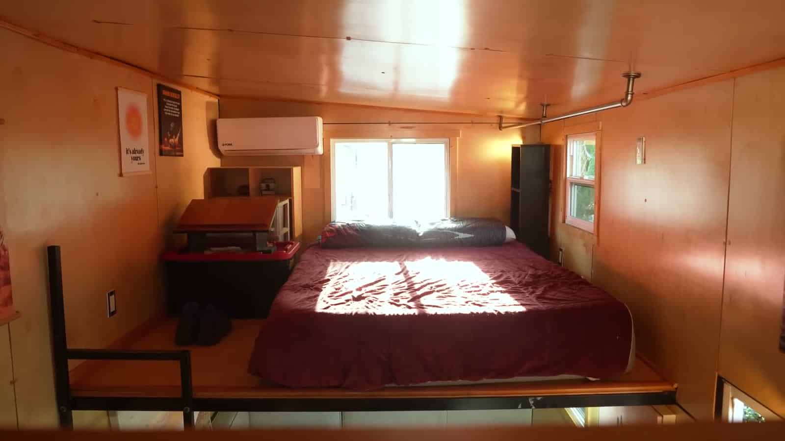 Primary sleeping loft of a tiny house with a king bed under a red cover, mini-split head, window at the gable end, and a black safety rail at the loft edge
