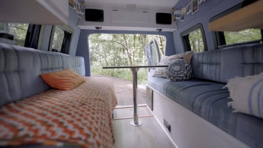 Interior of a retro van with cozy seating, a small table, and scenic forest views outside.