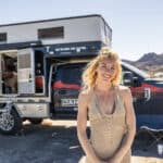 Woman smiling in front of a 4x4 truck camper in a desert setting, showcasing tiny living freedom.