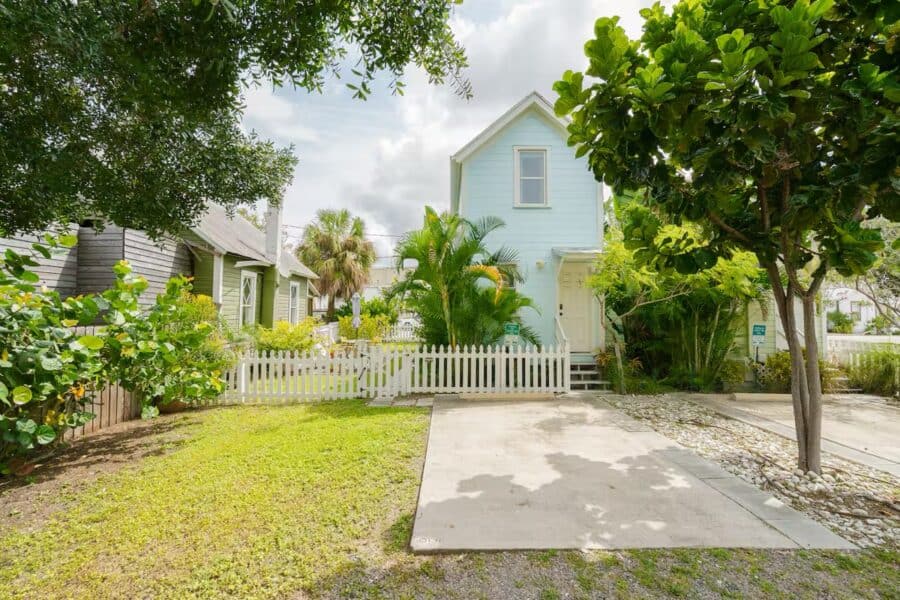 Bright blue tiny house surrounded by tropical trees and plants in a sunny Florida setting.