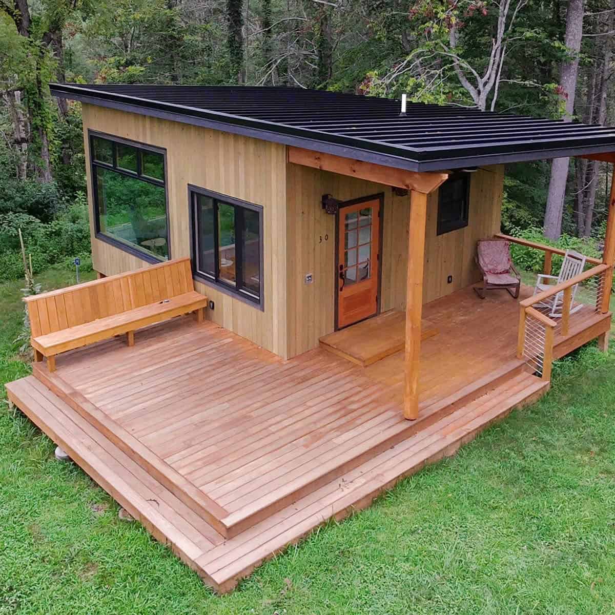 Compact tiny house with a spacious wooden deck in a forest setting.