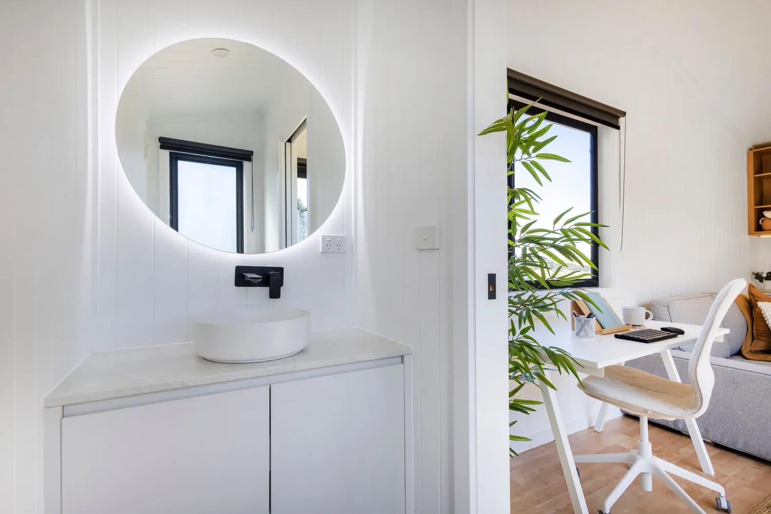 Bathroom vanity in the Joey tiny house with a backlit round mirror, white vessel basin on a stone-look benchtop, matte black wall-mounted mixer, white cabinetry, and a frosted window