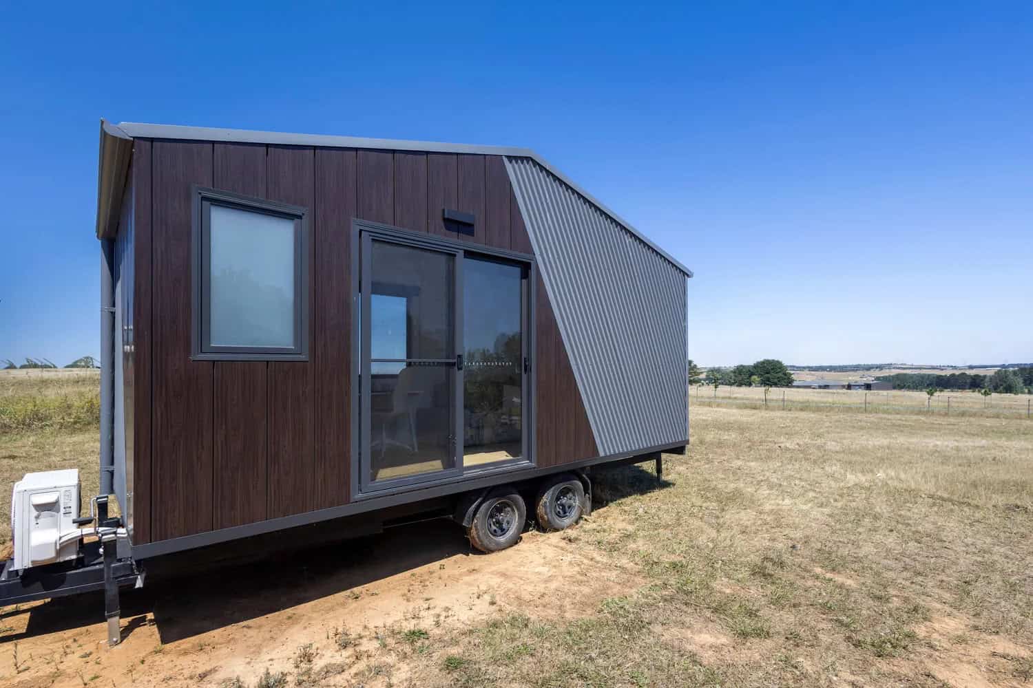 Joey tiny house by CozyCo with dark timber-look cladding, a diagonal corrugated Colorbond feature wall, sliding glass door, and dual-axle trailer, parked on dry Australian grassland
