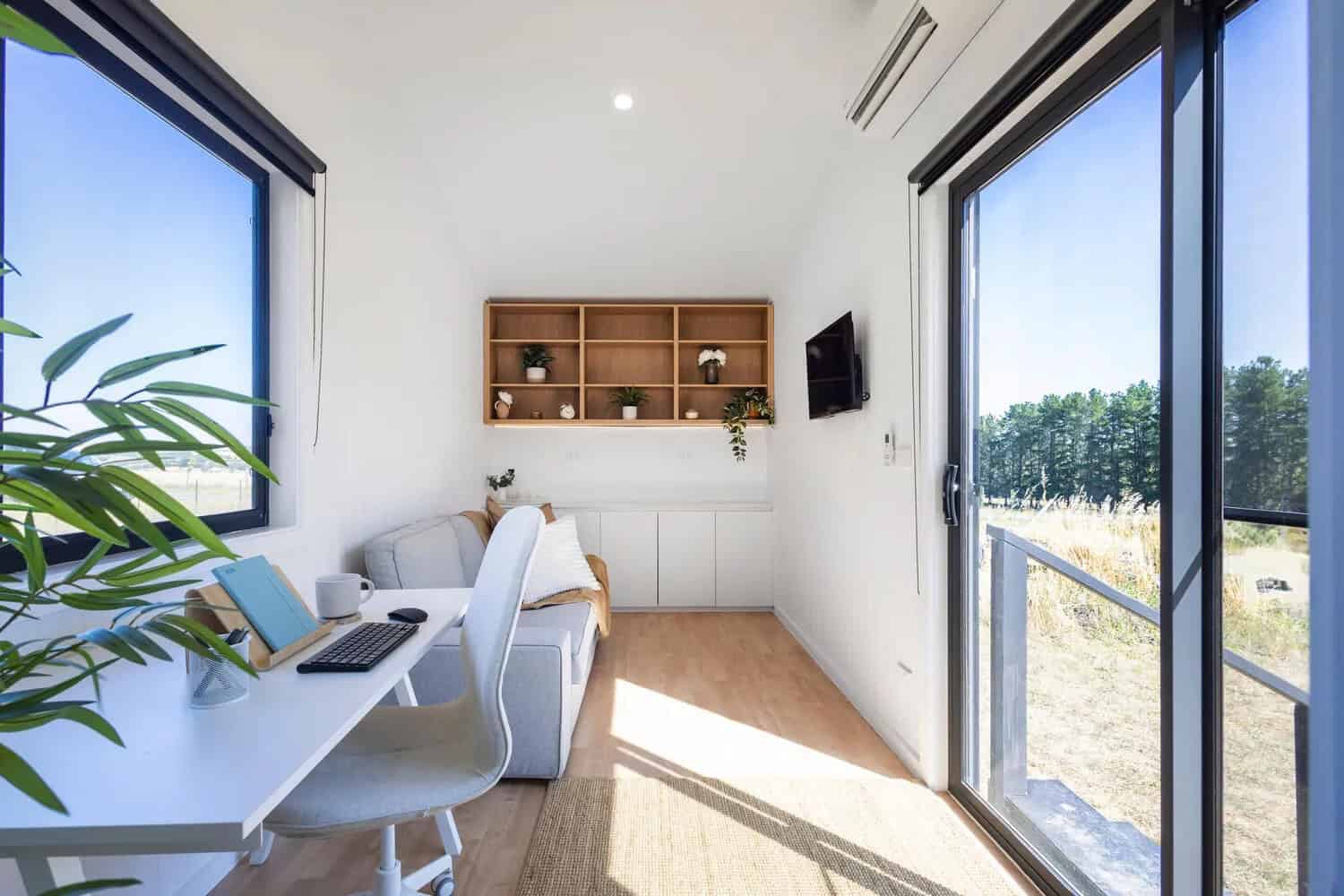 Interior of the Joey tiny house set up as a studio office, with a white desk under a window, grey sofa, white VJ paneling, timber open shelving, wall-mounted TV, split-system air conditioner, and sliding glass door