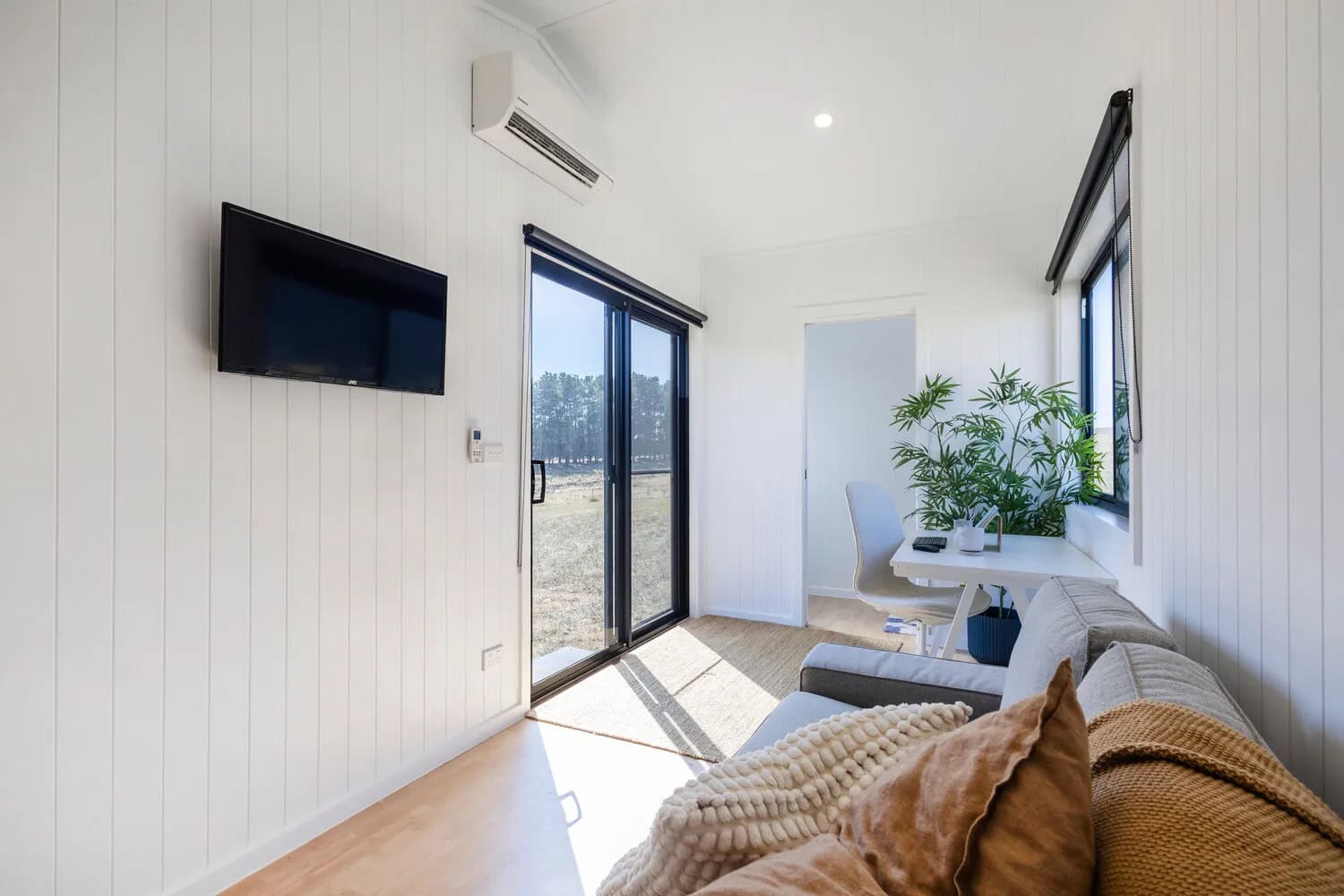 Living area of the Joey tiny house with a wall-mounted TV, split-system air conditioner, sliding glass door to the outside, desk and chair by the window, and the bathroom doorway beyond