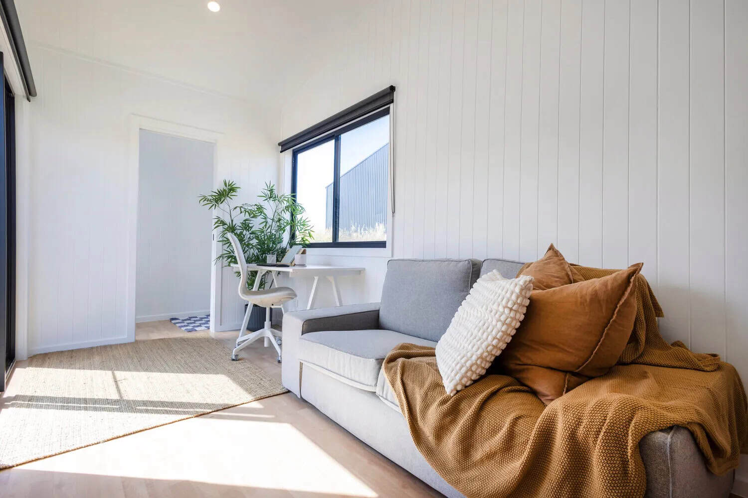 Interior of the Joey tiny house showing a grey sofa with cushions and throw, a white desk beneath a double-glazed window, jute rug, and the open doorway through to the bathroom