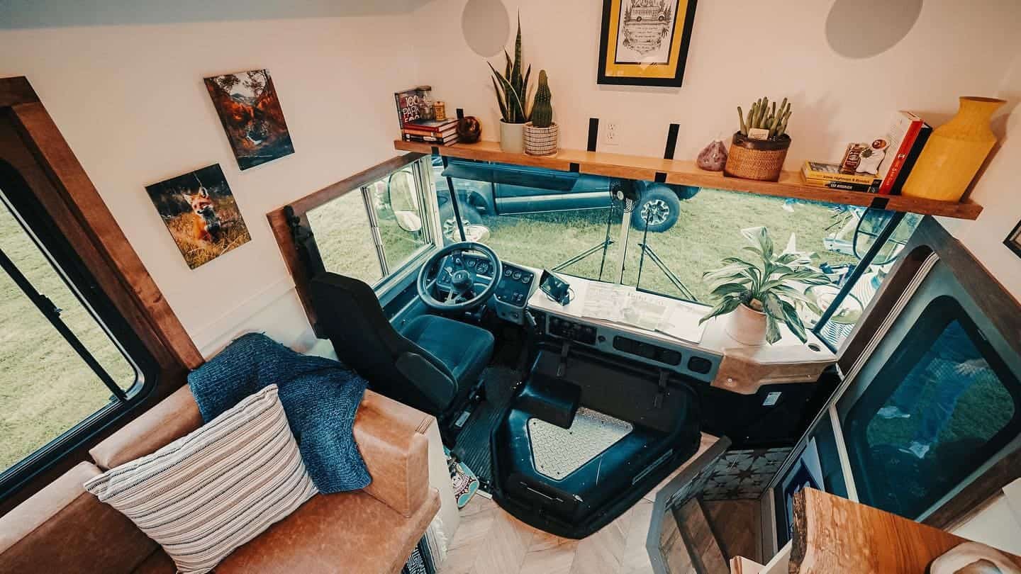 Front living area of the bus with a leather sofa, wood dash, houseplants, and a shelf above the windshield