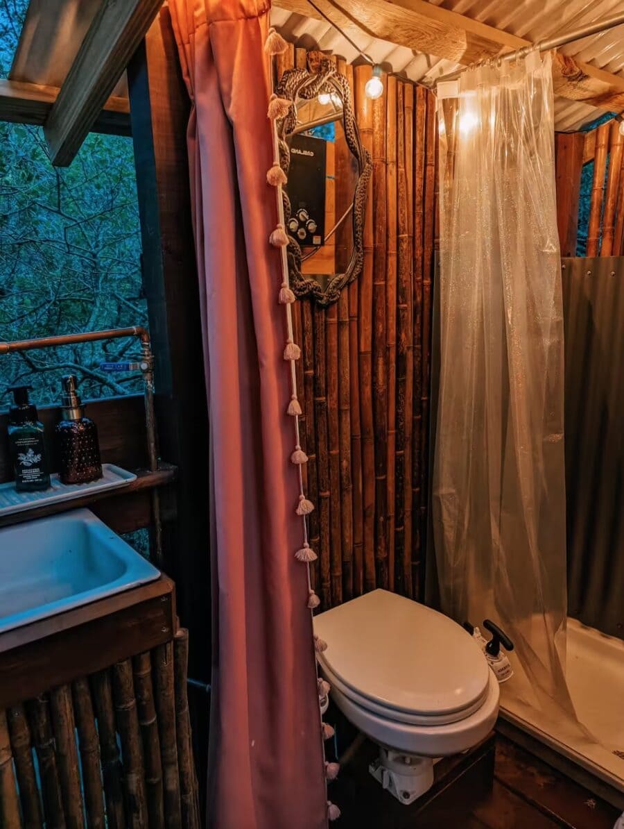 Rustic jungle bungalow bathroom with wooden walls, shower curtain, and scenic forest view.