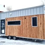 The Katrin tiny house by Craft House with wood and gray standing-seam siding, a gable roof, and a wood-stove chimney, in the snow