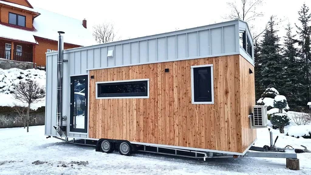 The Katrin tiny house by Craft House with wood and gray standing-seam siding, a gable roof, and a wood-stove chimney, in the snow