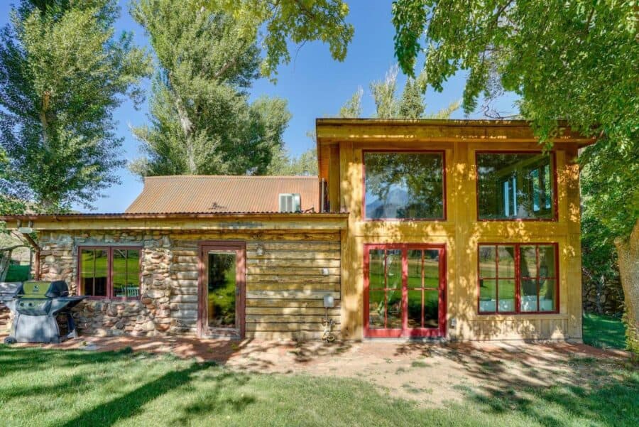 Log cabin with stone and wood exterior, surrounded by lush greenery and tall trees.