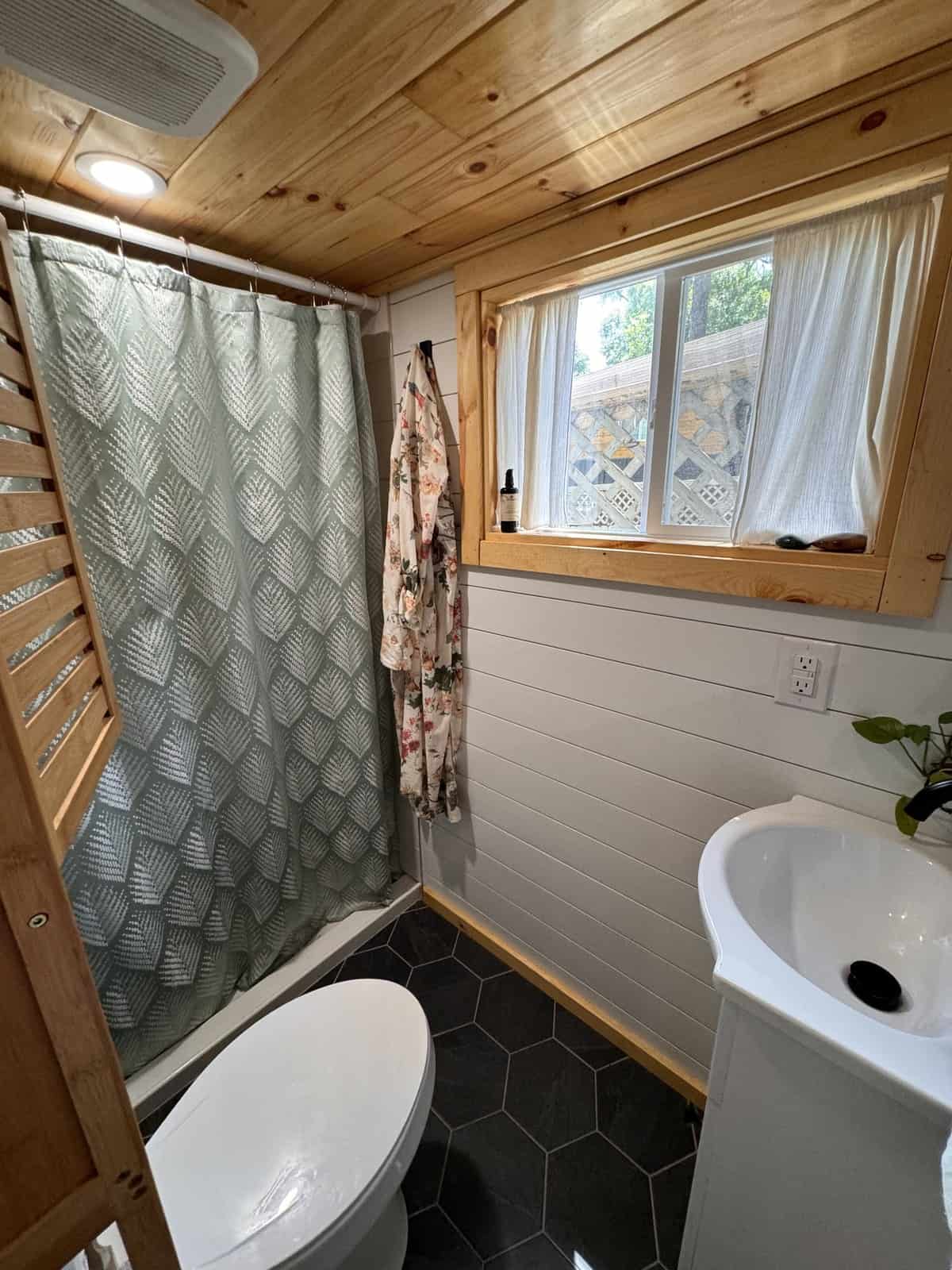 Full bathroom in the Leela tiny house with a curtained shower, flush toilet, vanity, black hex-tile floor, and a window