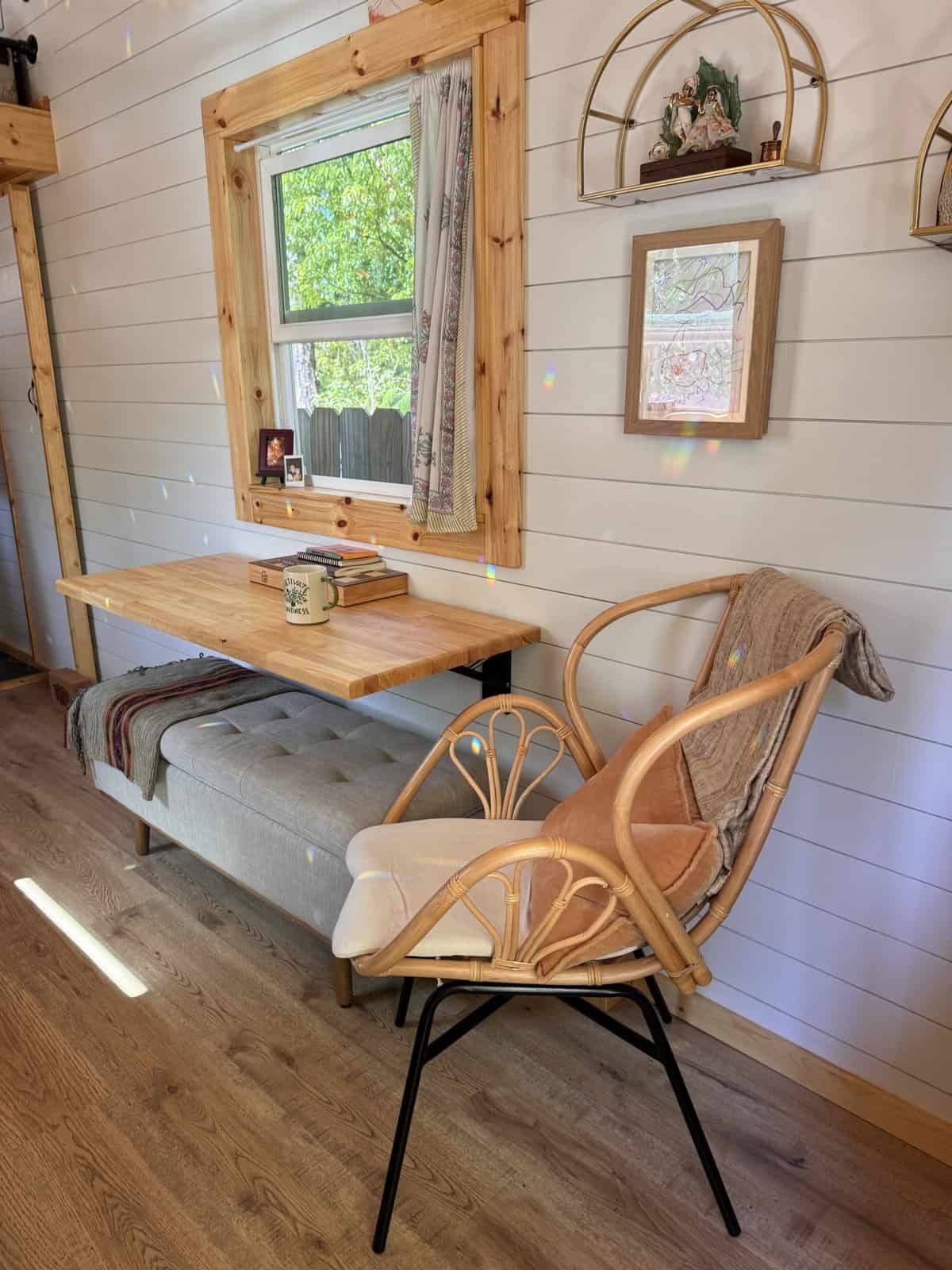 Fold-down butcher-block desk with a cushioned bench and a rattan chair beneath a window in the Leela tiny house