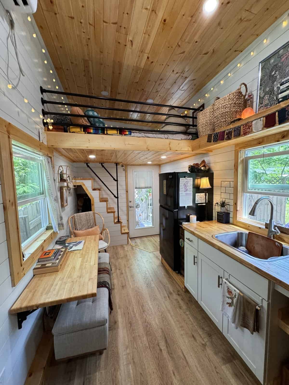 Wide view of the Leela tiny house with a string-lit loft and pipe railing, open stairs, a window-side desk and bench, and a butcher-block kitchen by the glass entry door