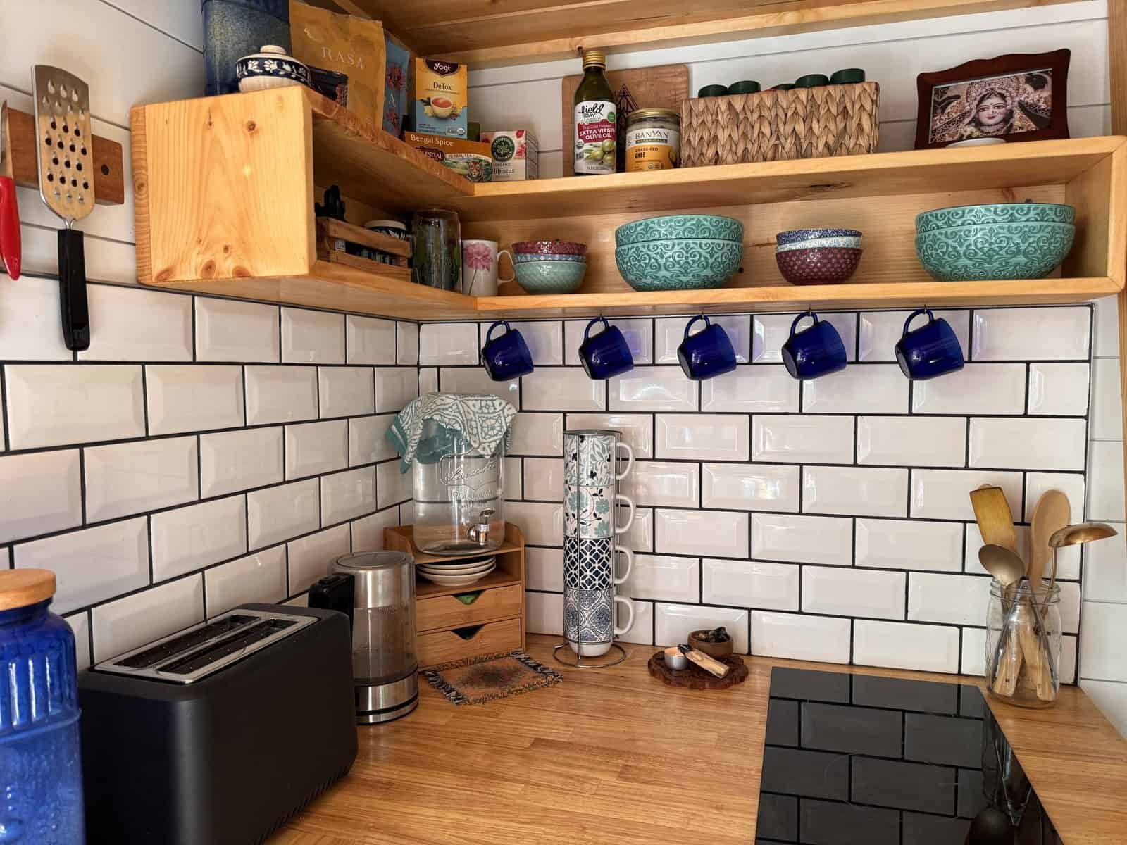 Leela tiny house kitchen with white subway tile, butcher-block counter, open corner shelving, mugs on hooks, and a toaster