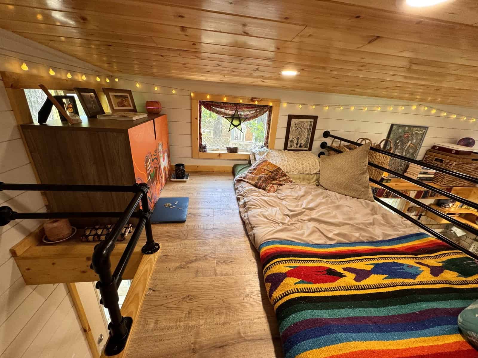 Master sleeping loft in the Leela tiny house with a bed, string lights, a dresser, and a window under the pine ceiling