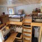 Interior of the Leela tiny house showing both sleeping lofts above a subway-tile kitchen, with a ladder, open stairs, and the bathroom below