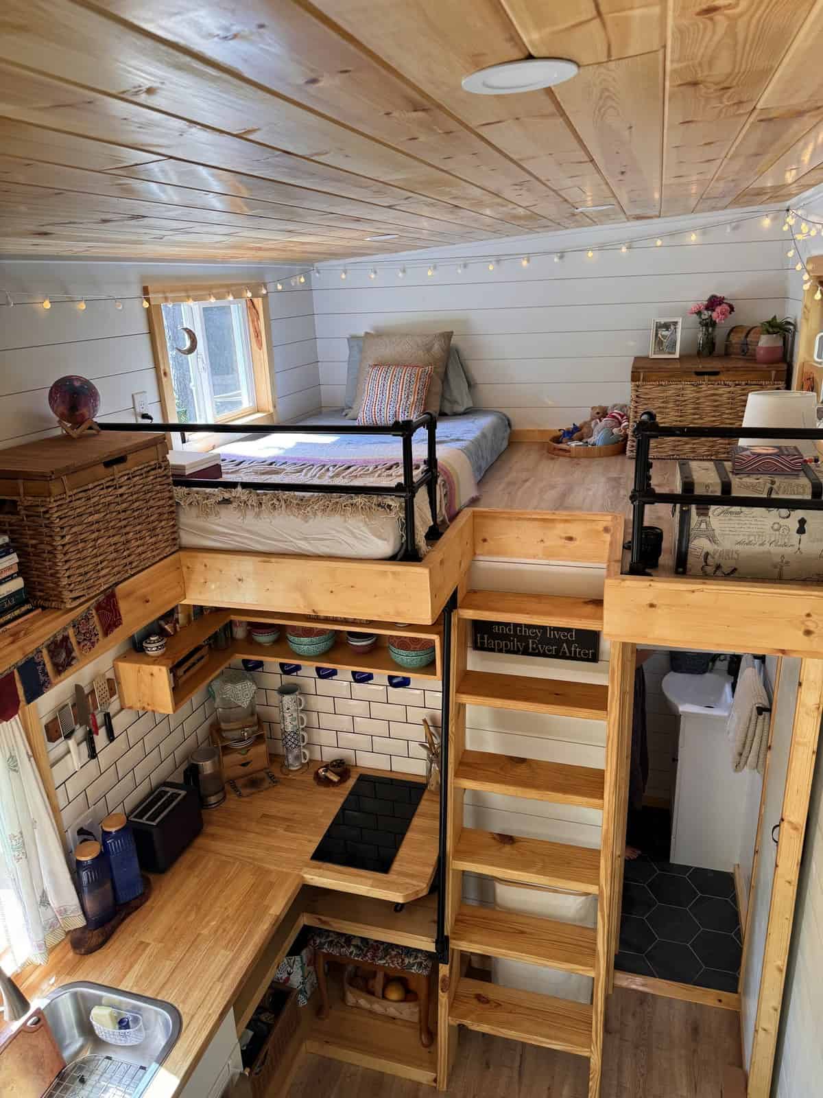 Interior of the Leela tiny house showing both sleeping lofts above a subway-tile kitchen, with a ladder, open stairs, and the bathroom below