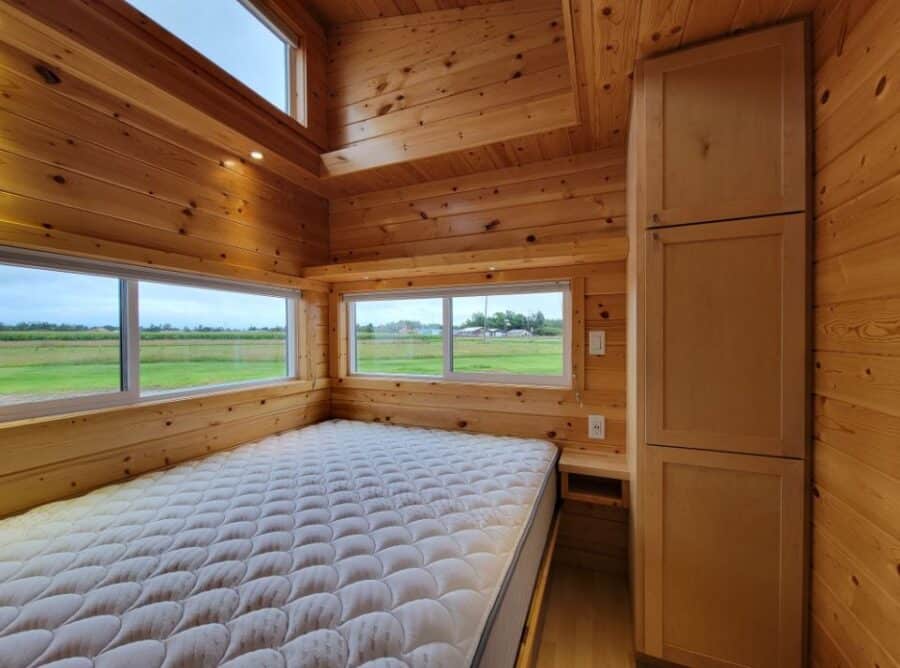 Compact tiny house bedroom with natural wood finishes and large windows.
