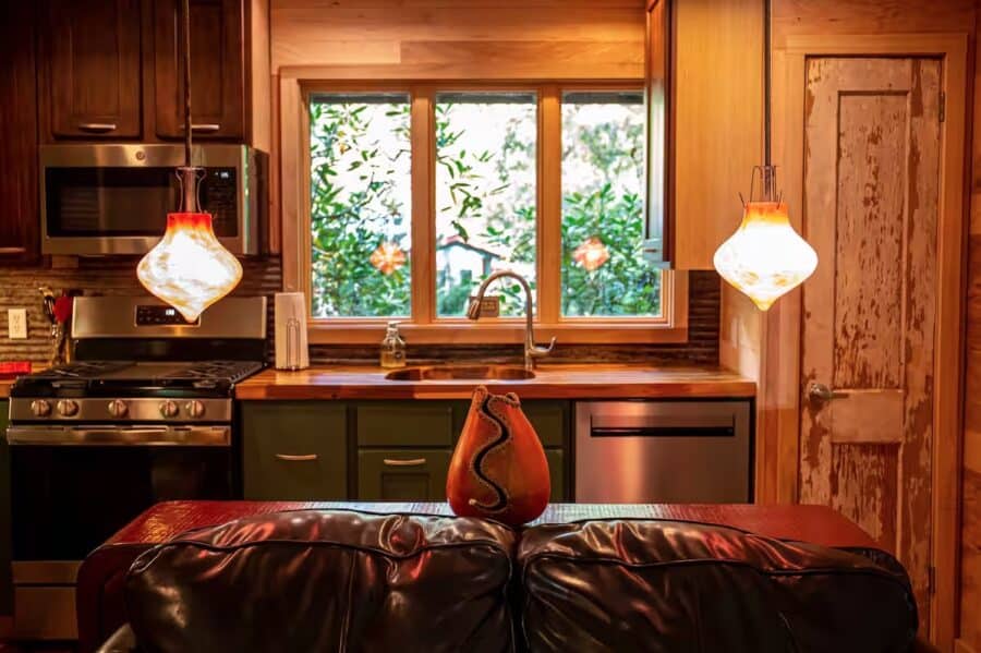 Charming tiny house kitchen with wooden cabinets, modern appliances, and a view of lush greenery thr.