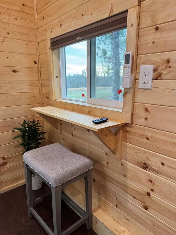Fold-down wood desk and stool under a window with a forest view inside the Mantra cabin, with the mini-split remote nearby