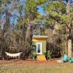 Lime-green Mantra micro cabin tiny house on wheels with a shed roof, hammock, picnic table, and teal chairs set among North Florida pines
