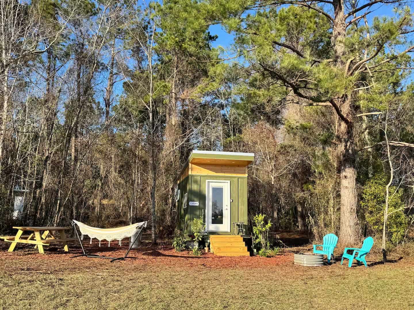 Lime-green Mantra micro cabin tiny house on wheels with a shed roof, hammock, picnic table, and teal chairs set among North Florida pines