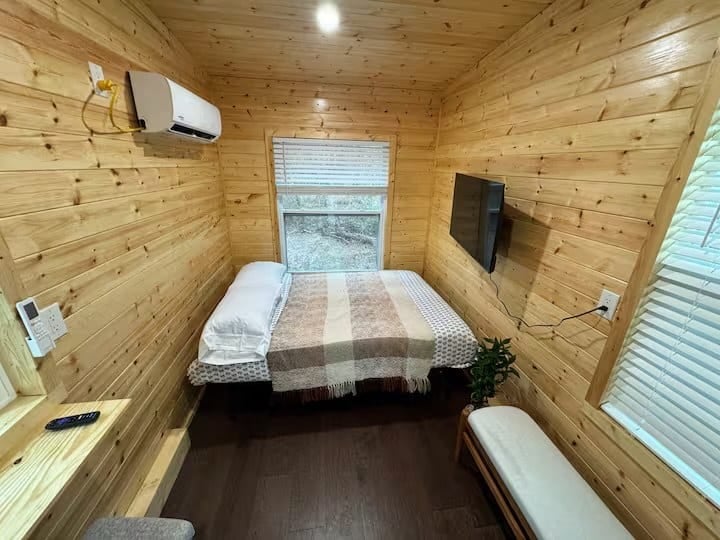 Interior of the Mantra glamping cabin showing the queen bed, wall-mounted mini-split, smart TV, fold-down desk, and pine walls