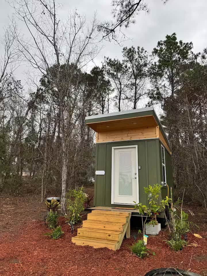 Olive-green Mantra micro cabin with a glass front door, cedar-trimmed shed roof, and wood steps surrounded by mulched garden beds