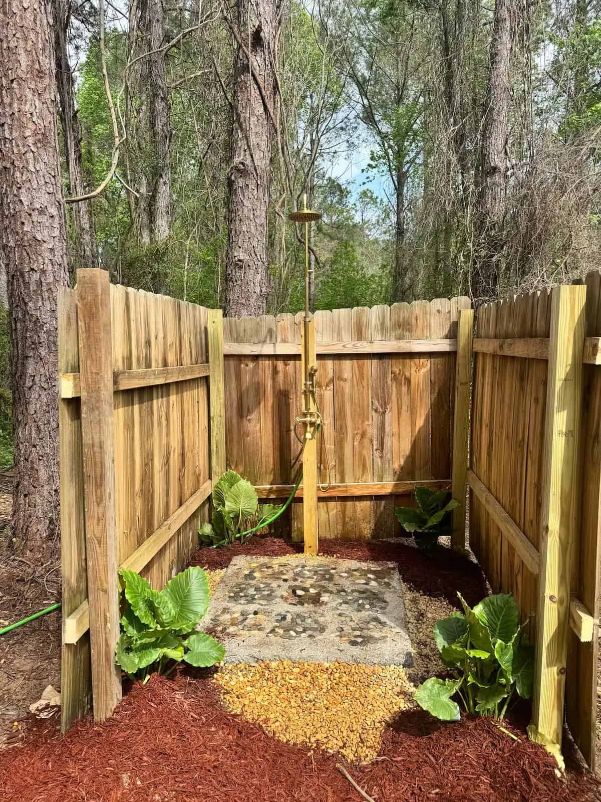 Wood-fenced outdoor shower with a pebble stone floor and tropical plants among the trees