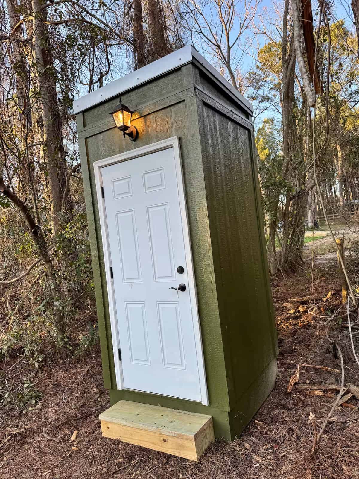 Green wood outhouse with a white door and a lantern, tucked into the woods across from the cabin