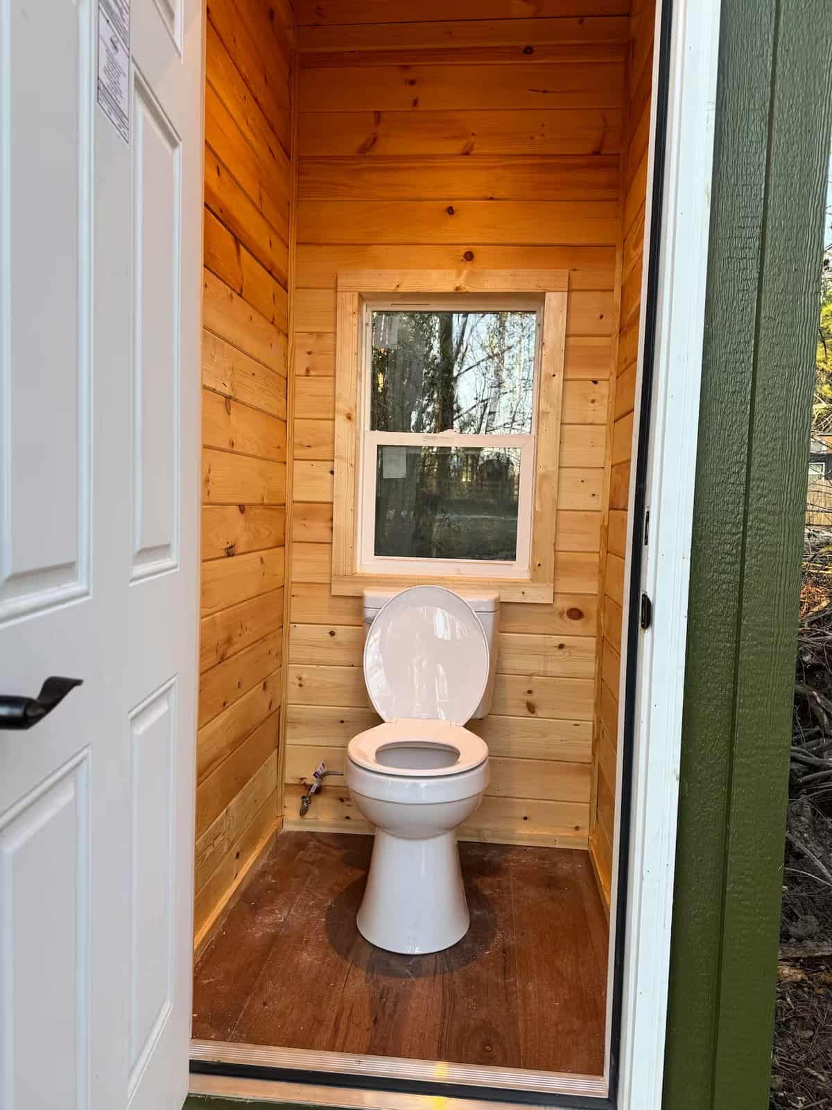 Inside the shared outhouse: a full flush toilet with pine tongue-and-groove walls and a window to the trees
