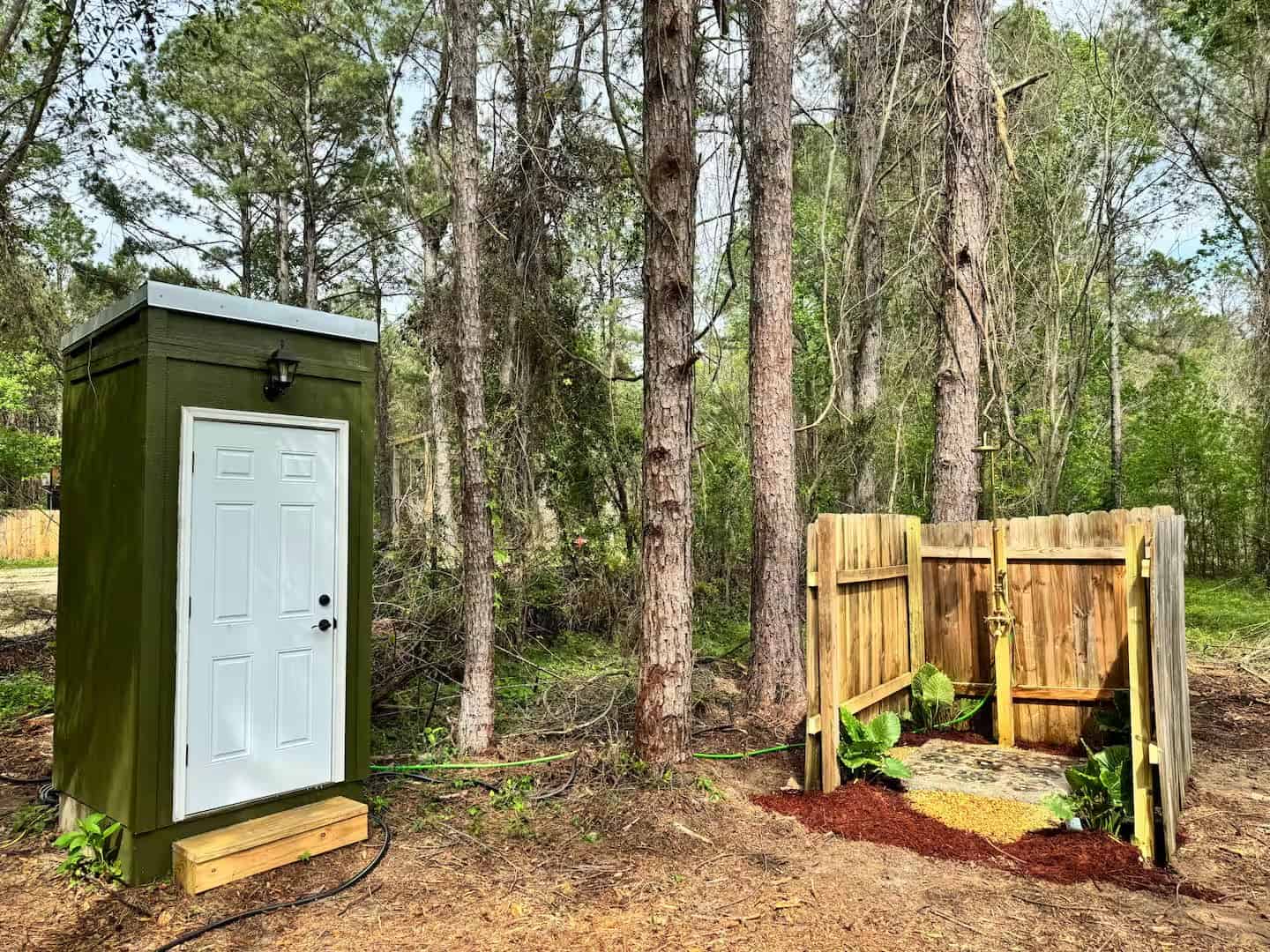 Green outhouse and a wood-fenced outdoor shower set among the pines near the Mantra glamping cabin
