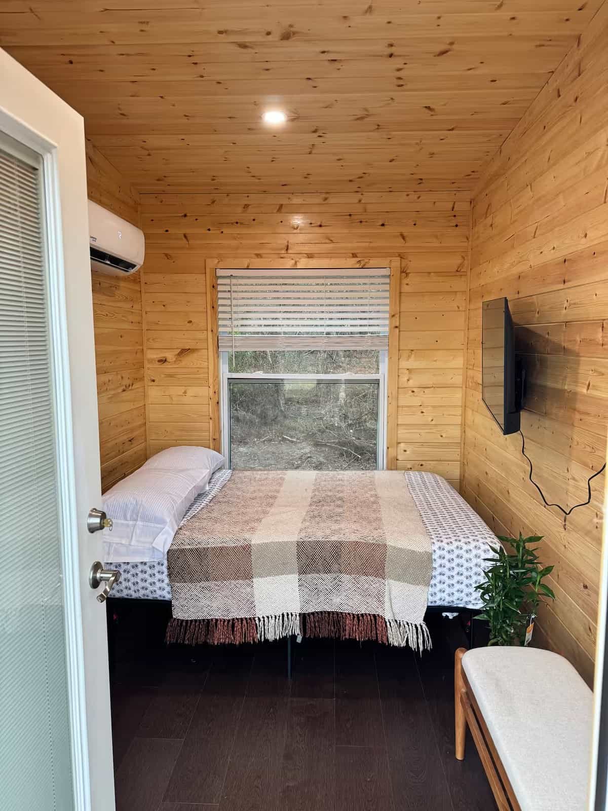 Queen memory-foam bed in the Mantra cabin beneath a large window looking out into the trees, with a mini-split and smart TV