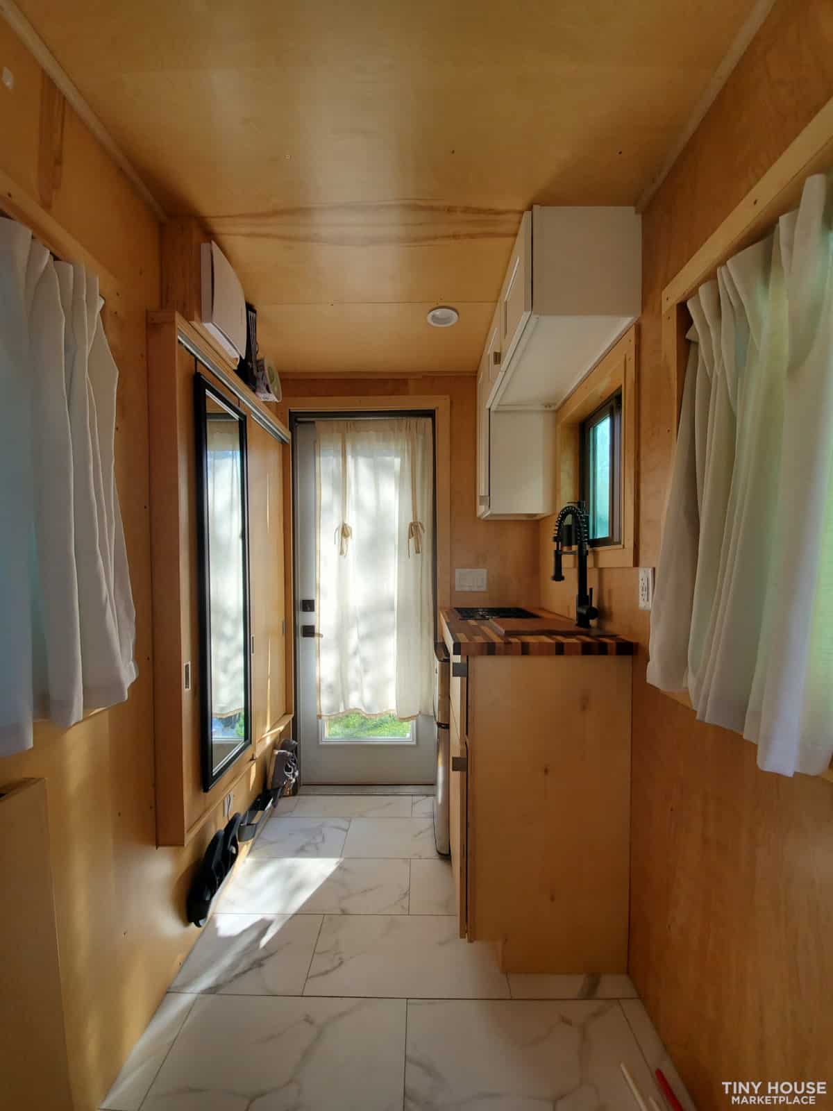 Interior view of a compact tiny house kitchen with wooden walls and white cabinetry.