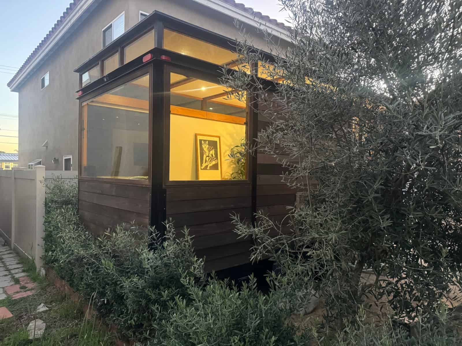 Corner glass wall of the MoD tiny house glowing at dusk beside olive trees