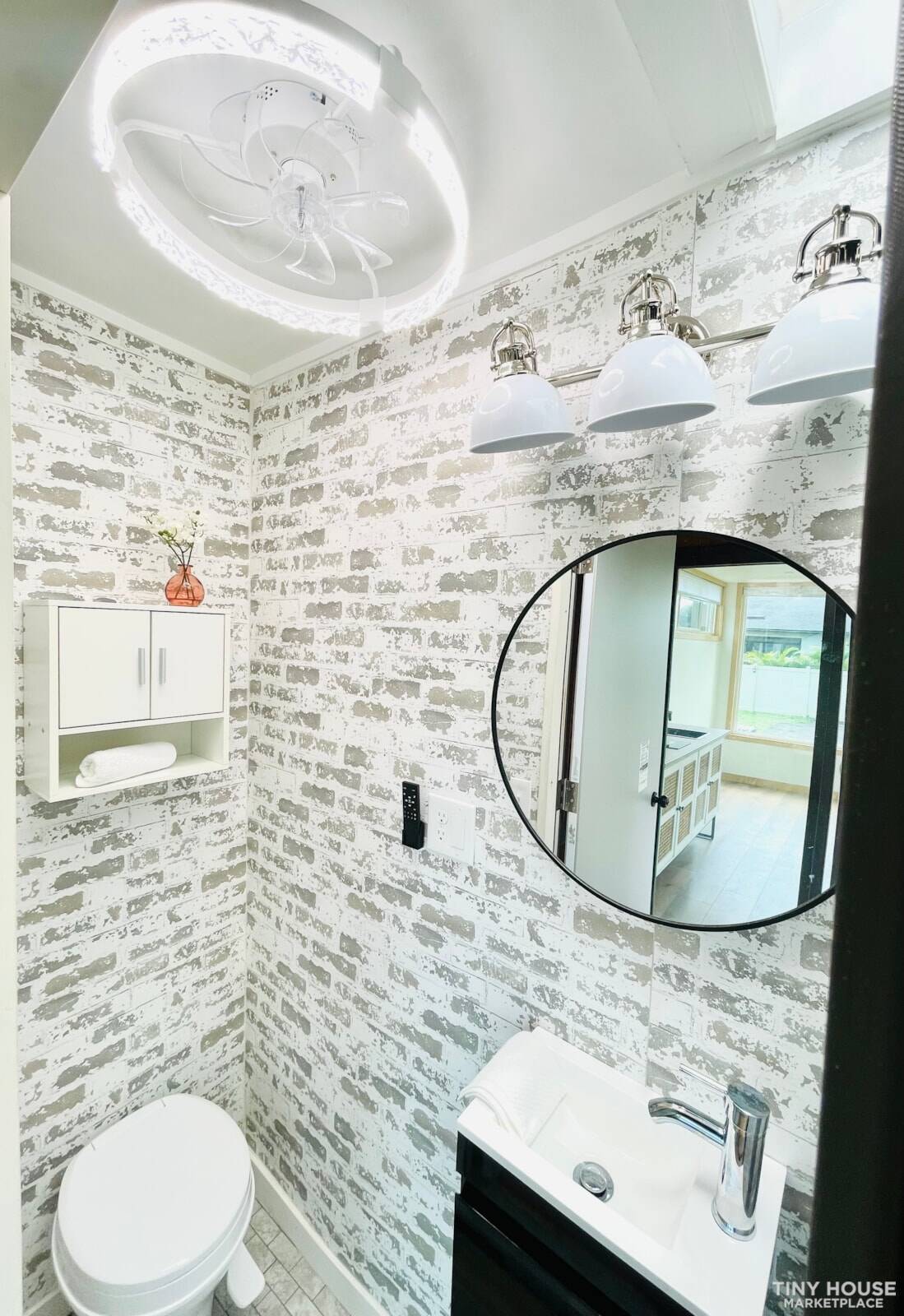 Industrial-chic bathroom with distressed brick walls, floating black vanity, round mirror, LED ceiling fan, and skylight