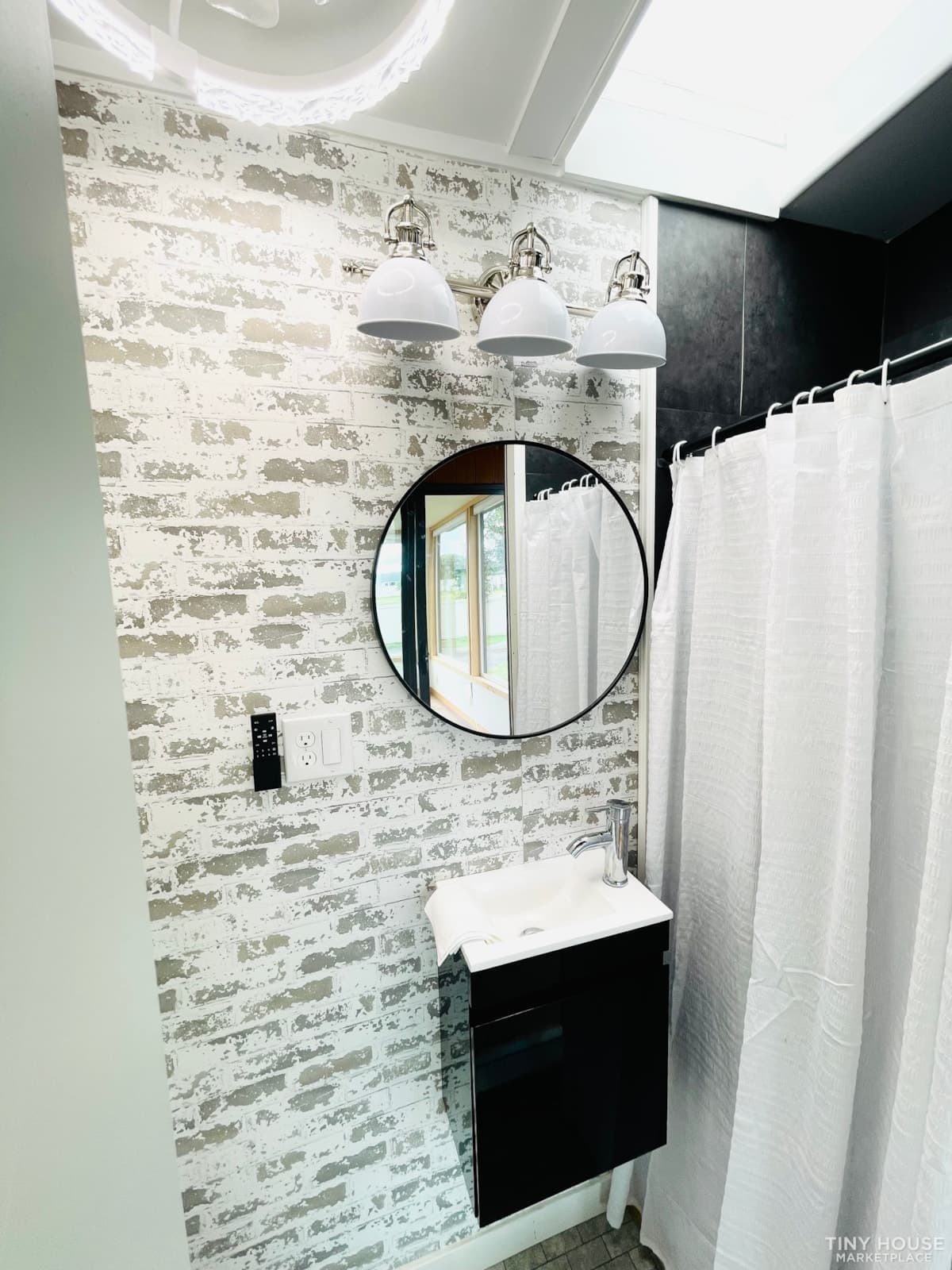 Close-up of bathroom vanity with distressed white brick walls, round mirror, dual sconce lighting, and skylight