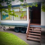 Tiny house on wheels with wooden stairs and large windows.
